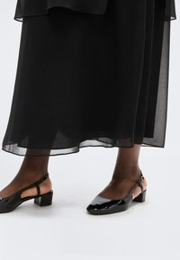 Σκούρα πόδια με μαύρα παπούτσια slingback από δερμάτινο γυαλιστερό με χαμηλό τακούνι κάτω από μια διάφαν η μαύρη μέση φούστα, που στέκονται σε λευκό πάτωμα.