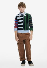 Mehrfarbiger Strickpullover mit Streifen und Mustern, kombiniert mit braunen Cargo-Hosen. Accessoires sind ein weißes Bandana und schwarze Slippers.