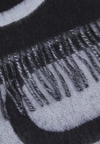 Vilnonis šalikas su juodos ir pilkos spalvų scheme, turintis modernų abstraktų raštą; apipjaustytos kraštinės su kutais, suteikiančiais tekstūrą ir detalumą.