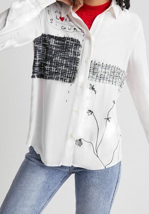 Bílá košile na knoflíky s černo-bílými tweedovými záplaty, černými květinovými kresbami a textem s červeným srdcem, nošená přes červené tričko a modré džíny.