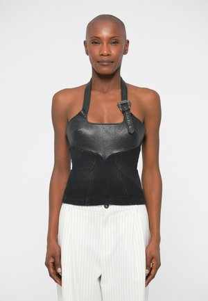 Mulher com cabeça rapada, vestindo um top halter de couro preto com fivela e calças brancas com riscas verticais, em pé contra um fundo neutro.