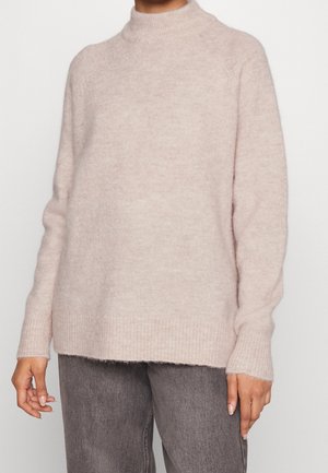 Lichtroze, fuzzy gebreide trui met een hoge halslijn, geribde manchetten en zoom. Heeft een ontspannen pasvorm en een zachte textuur. Draagt over grijze jeans.