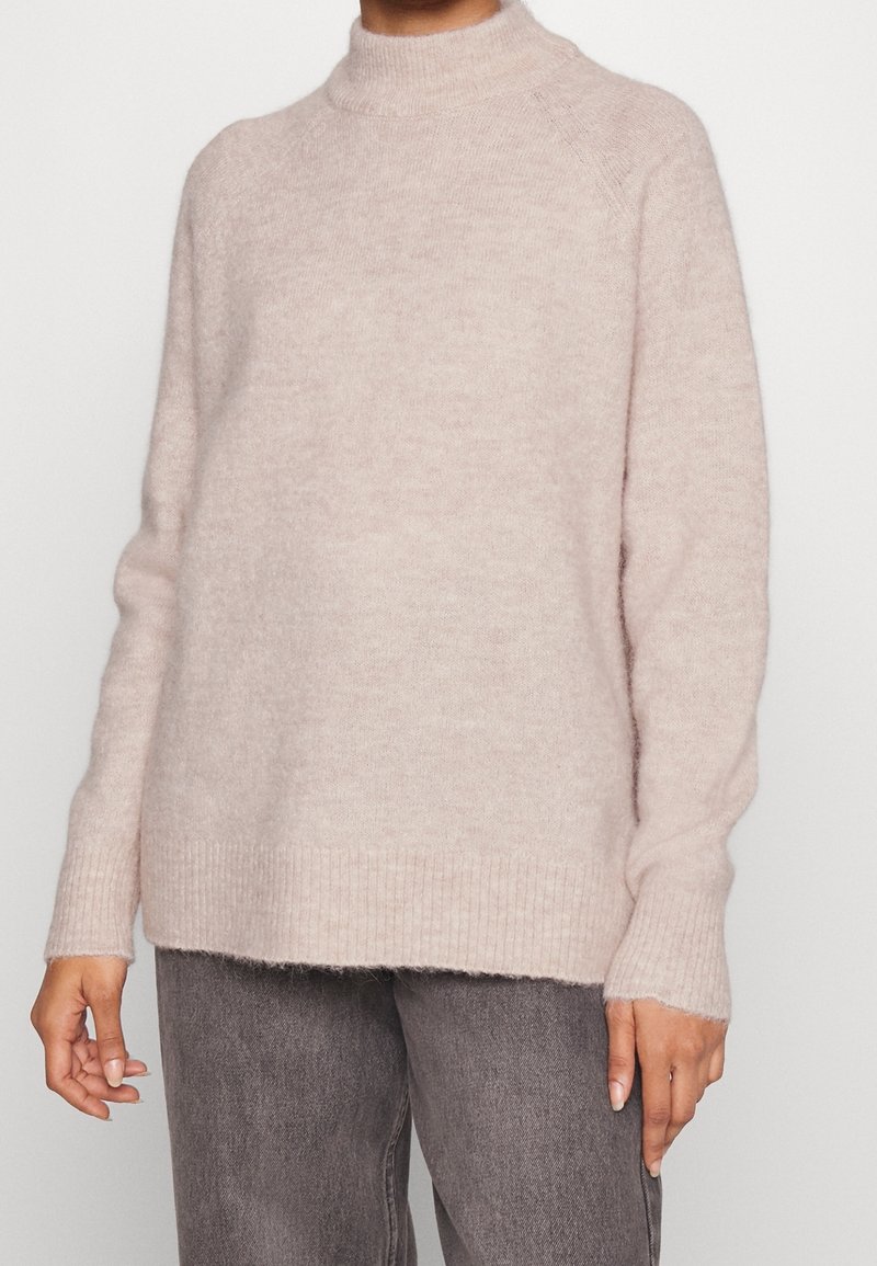 Ljusrosa, fluffig stickad tröja med hög krage, ribbade ärm- och nederkanter. Har en avslappnad passform och mjuk textur. Bärs över grå jeans.