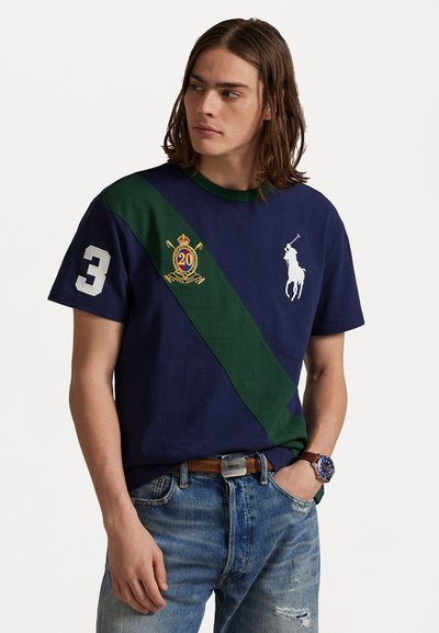 Granatowy T-shirt z krótkim rękawem z zielonym ukośnym pasem, białym logo polo, numerem 3 na rękawie i haftowanym herbem. Materiał bawełniany.