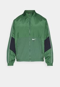 Zelená nylonová větrovka s vysokým límcem, zipovým zapínáním a námořnickými akcenty na bocích. Obsahuje bílé logo Nike a strukturované panely.