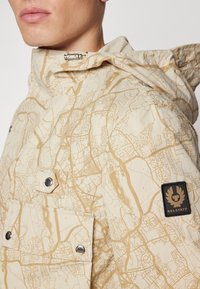 Beige jacka med karttryck, silverdetaljer och en svart Belstaff-logotillbehör. Huva och tryckknappsöppning, samt fickdetalj.