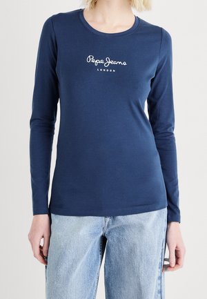 Femme portant un t-shirt à manches longues bleu marine Pepe Jeans London et un jean bleu clair, debout devant un fond clair uni.