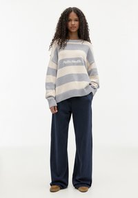 Maglione a righe in beige chiaro e grigio, con la scritta "Pacific Republic" al centro, abbinato a pantaloni larghi blu navy e calzature marroni.