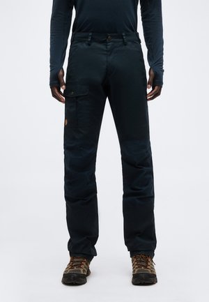 Άτομο που φοράει μαύρο παντελόνι τύπου cargo με τσεπάκι με καπάκι στον μηρό και καφέ παπούτσια πεζοπορίας, στέκεται μπροστά σε λευκό φόντο.