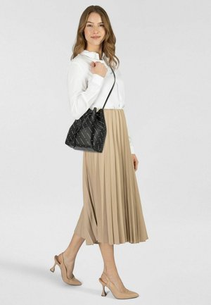 Schwarze Bucket Bag mit weißem Schriftmuster, quer über die Schulter getragen. Model in einem weißen Hemd und einem beigefarbenen Faltenrock, kombiniert mit nude hohen Schuhen.