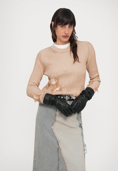 Femeie într-un pulover bej cu model striat și uzat, fustă din denim bicolor, curea neagră cu ținte și mănuși negre din piele, pozând pe fundal alb simplu.