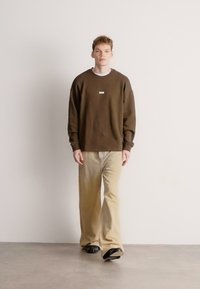 Felpa marrone con scollo rotondo e piccola toppa con logo, abbinata a pantaloni beige chiaro a gamba larga e scarpe nere, su sfondo neutro.