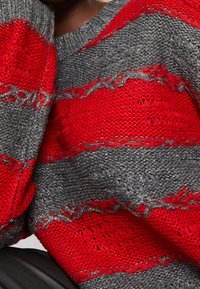 Närbild av en stickad tröja med breda röda och grå ränder som har ett texturerat, något slitet mönster.