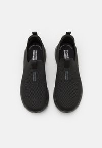 Zapatos negros tipo slip-on con parte superior de malla, suela flexible y tacón plano. Presentan una pequeña pestaña para facilitar el calzado y una marca en los costados.
