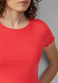 Vrouw in een aansluitend, rood, geribbeld T-shirt met korte mouwen en een gekartelde rand op de mouwen, staand tegen een grijze achtergrond.