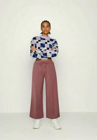 Mulher de pé com os braços cruzados, vestindo um suéter xadrez azul, preto e cinza, calças largas em rosa mauve e ténis brancos, contra um fundo simples.