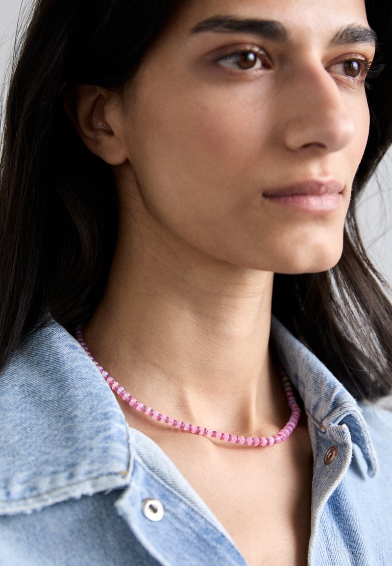 Ružový korálkový náhrdelník s malými, zaoblenými korálkami, ktorý obsahuje zmes svetloružových a tmavoružových odtieňov, nosený cez svetlo denimovú košeľu.