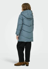Padded winter coat in soft blue with a hood, featuring chevron stitching details. Worn with black, loose-fit trousers and brown ankle boots.