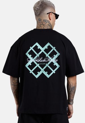 Person mit kurzen grauen Haaren und Nacken-Tattoo, die ein schwarzes T-Shirt mit großem blauem geometrischem Muster und kursivem Text auf dem Rücken trägt.