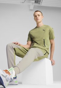 Svetle zelené sportovní tričko a béžové kalhoty s černými detaily a kapsou. Model má na sobě bílé tenisky s barevnými prvky.