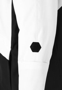 Weiße und schwarze Jacke mit einem hexagonalen schwarzen Stoffpatch am Ärmel. Der Stoff wirkt glatt und hat ein sauberes Design.