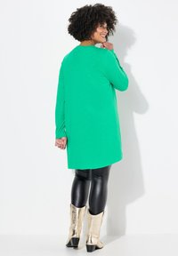 Grünes langärmliges Tunika mit abgerundetem Saum, kombiniert mit schwarzen Leggings und metallisch goldenen Stiefeletten. Strukturierter Stoff, lockere Passform.