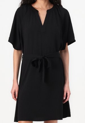 Woman wearing a short-sleeve black dress with a V-neck and a tied belt at the waist, standing against a plain white background.