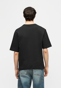 Άνδρας με απλό μαύρο T-shirt με κοντά μανίκια και ξεθωριασμένο μπλε τζιν, φαίνεται από πίσω με λευκό φόντο.