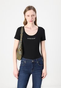 Černý krátký rukáv bodysuit s bílým potiskem "GUESS JEANS", kombinovaný s modrými džíny a olivově zelenou křížovou taškou.