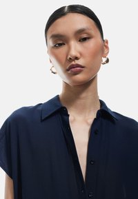 Camicia blu navy con colletto, tessuto leggero e maniche corte. Il modello indossa orecchini a cerchio dorati. Dettagli della superficie texturizzati visibili.