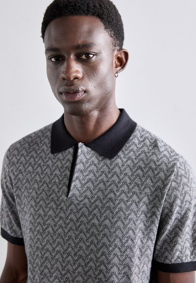 Polo en noir et blanc avec un motif en zigzag, doté d'un col et de poignets noirs unis. Texture douce, manches courtes et sans fermetures.