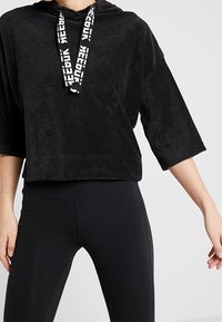 Sweat à capuche court en peluche noire avec un cordon de serrage et un logo blanc. Il a des manches courtes et une coupe décontractée, associé à des leggings noirs.