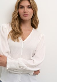 Blusa bianca a trama con scollatura a V, maniche a palloncino e chiusure con bottoni. Il tessuto presenta un motivo stropicciato e una consistenza morbida e fluida.