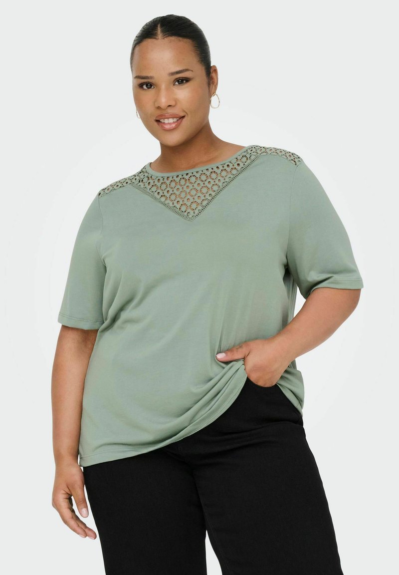 T-shirt verde a maniche corte con un motivo in pizzo sul collo, vestibilità rilassata, tessuto morbido e tasca laterale. Abbinato a pantaloni neri.