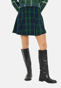 Beine, die schwarze knöchelhohe Lederstiefel und einen plissierten Karorock in Grün und Marineblau tragen, mit einem teilweise sichtbaren grünen Pullover.