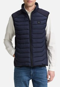 Doudoune sans manches bleu marine avec un col montant et une fermeture éclair à l'avant. Présente un motif matelassé horizontal et des poches latérales. Portée par-dessus une chemise claire.