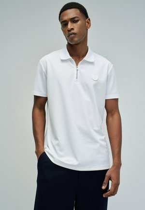 Jeune homme portant un polo blanc à manches courtes avec une fermeture éclair et un logo rond, debout devant un fond gris uni.