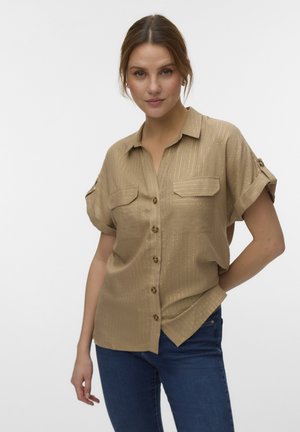 Vrouw draagt een beige korte mouwen blouse met knopen en gouden strepen en blauwe jeans, staand met één hand in haar zak.
