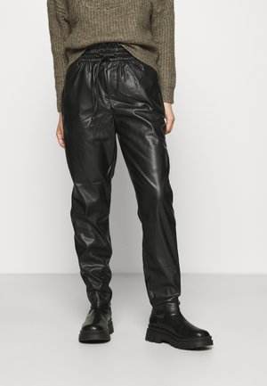 Person iført sorte joggerbukser i kunstlæder, chunky sorte støvler og en brun striktrøje, stående foran en ensfarvet hvid baggrund.