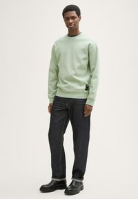 Sudadera verde clara, con cuello de canalé, etiqueta de logo prominente en el lado. Llevada con jeans de mezclilla oscura y botas de cuero negras, con los puños doblados.
