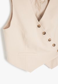 Gilet beige en matière lisse, avec un décolleté en V, cinq boutons marron et deux poches latérales. Forme compacte et ajustée.