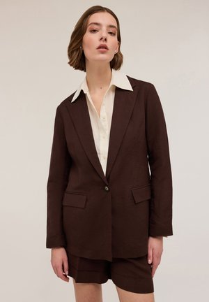 Bruine linnen blazer met patchzakken en een enkele knoop, gecombineerd met een crèmekleurig overhemd met kraag. Korte bruine shorts maken de outfit compleet.