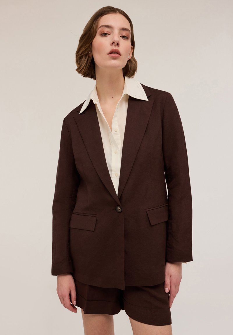 Brun linneblazer med fickor och en knapp, i kombination med en krämfärgad skjorta med krage. Korta bruna shorts kompletterar outfiten.