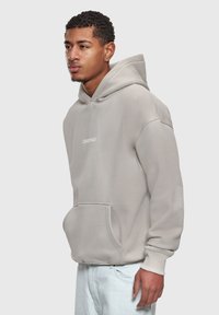 Joven con cabello rizado corto que lleva una sudadera con capucha beige claro con bolsillo frontal y pantalones blancos, de pie contra un fondo liso.