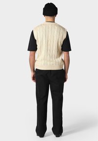 Béžový svetr bez rukávů s pleteným vzorem přes černou košili s krátkým rukávem, doplněný černými kalhotami; minimalistický design s žebrovaným lemem.