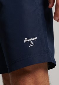 Costume da bagno corto blu navy in materiale leggero, con logo "Superdry Co" ricamato in bianco sull'orlo e cuciture ordinate.
