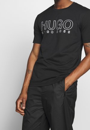 Homme portant un t-shirt noir Hugo Boss avec un logo blanc, un pantalon noir avec une poche zippée et un tatouage de diamant sur l'avant-bras gauche.