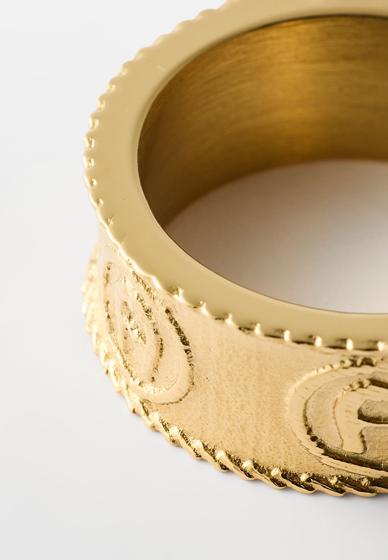 Goldfarbener Metallarmreif mit strukturierter Oberfläche, über das sich eine gewellte Kante und geprägte Muster erstrecken, die ein glänzendes Finish aufweisen.