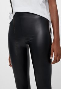 Svarta leggings i konstläder har en slank design med hög midja, mjuk yta och figurnära silhuett, i kombination med en vit topp.