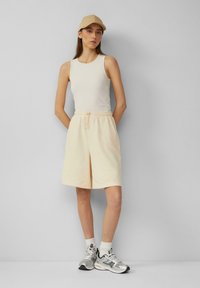 Beige geribbelde tanktop gecombineerd met losse lichte shorts. Witte sokken en zilveren sneakers maken de look af, tegen een grijze achtergrond.
